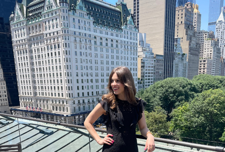 White girl with brown hair standing on a rooftop wearing a black dress in front of The Plaza