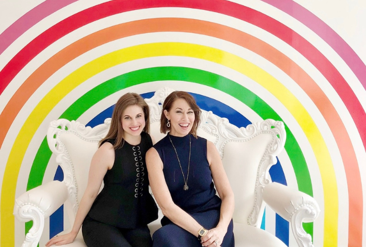 two white women with brown hair sit on a white chair in front of a rainbow mural