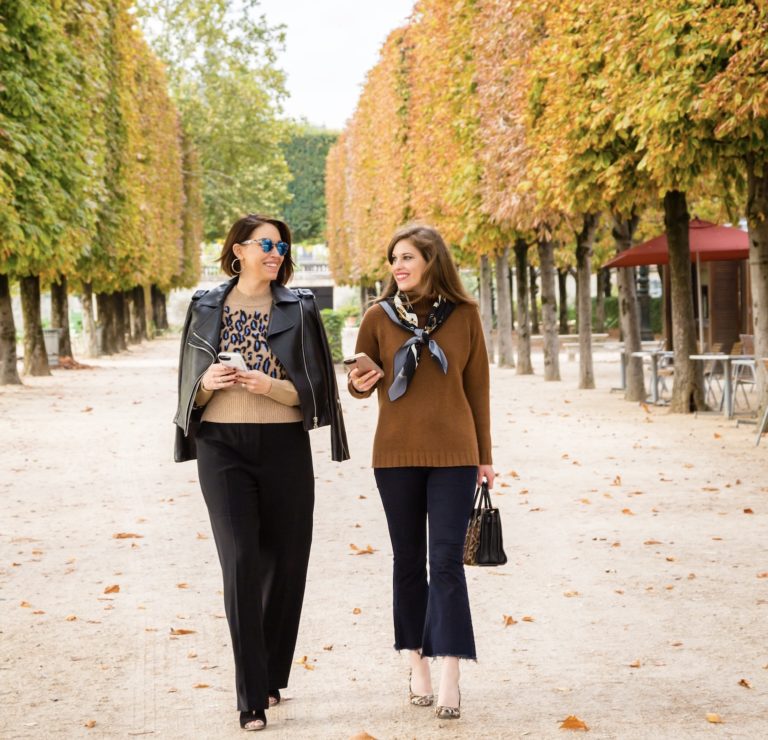 Two white women with brown hair wearing brown sweaters walking in Paris