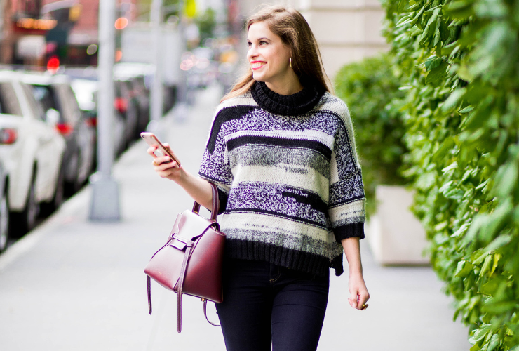 White girl with brown hair walks down the street & wears a multi-colored black & white sweater