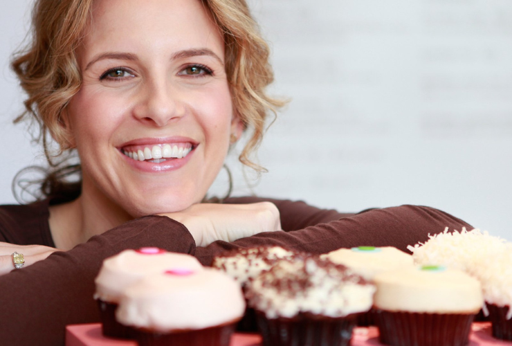 White woman with light brown hair smiles at the camera in front of cupcakes
