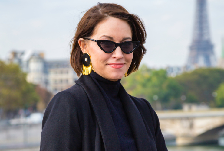 woman with brown hair wearing black sunglasses and black coat and statement earrings in front of Eiffel Tower