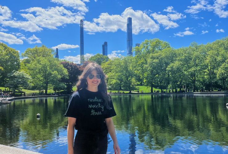 White woman with brown hair wear a black tee and sunglasses walking in front of a pond