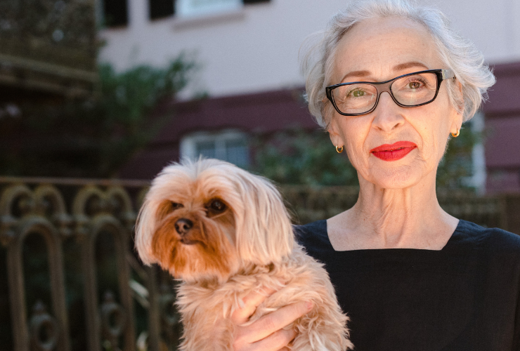 White woman with silver hear wearing red lipstick & black glasses holding a dog wearing a black top