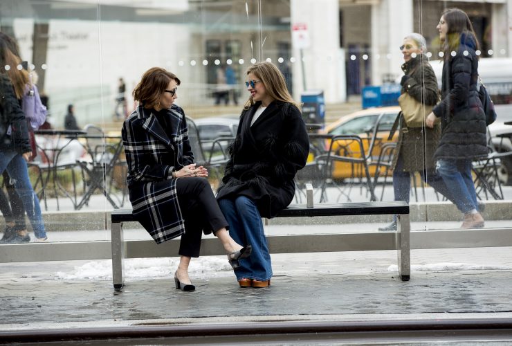 Two women sitting at a bus stop: one wearing a plaid coat, one wearing a blue coat. Both in sunglasses and heels.